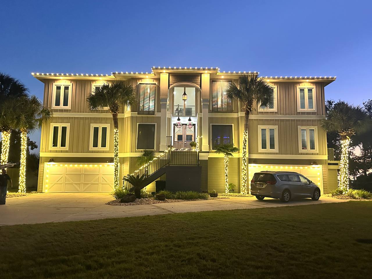 Multi-level Gulf Breeze home with warm-white roofline lights and wrapped palm trees.